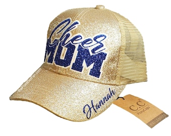 May include: Gold glitter trucker hat with blue glitter lettering that says "Cheer Mom" and the name "Hannah" on the brim. The hat has a mesh back and a brown tag that says "C.C. Pony Caps Exclusives".