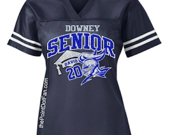 May include: A navy blue jersey with white stripes on the sleeves. The jersey has a white and blue glitter design that says "Downey Senior" with a graduation cap and a knight's helmet. The number "20" is also included in the design.