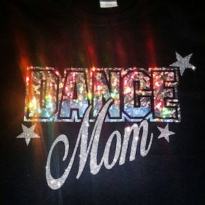 Può includere: Maglietta nera con lettere di glitter argentate che recitano "Dance Mom" con due stelle di glitter argentate su entrambi i lati del testo.
