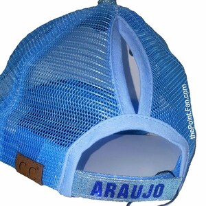 May include: Light blue trucker hat with a mesh back and a ponytail opening. The hat features the name "ARAUJO" in blue glitter lettering on the adjustable strap. A small brown leather patch with "CC" is on the side.