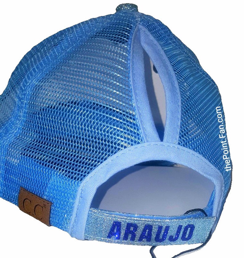 May include: Light blue trucker hat with a mesh back and a ponytail opening. The hat features the name "ARAUJO" in blue glitter lettering on the adjustable strap. A small brown leather patch with "CC" is on the side.