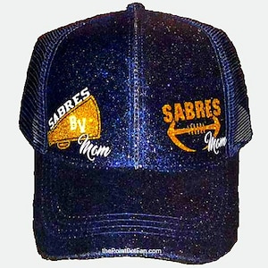 May include: A navy blue glitter trucker hat with the word "SABRES" in gold lettering, along with a megaphone and football graphic. The text "By Mom" is below the megaphone and football. The hat has a mesh back and a curved brim.