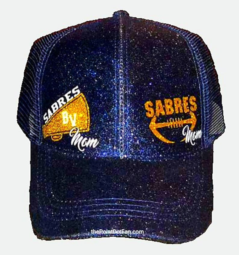 May include: A navy blue glitter trucker hat with the word "SABRES" in gold lettering, along with a megaphone and football graphic. The text "By Mom" is below the megaphone and football. The hat has a mesh back and a curved brim.