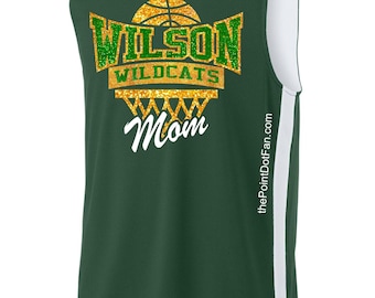 May include: Green basketball jersey with white trim and a V-neck. The front features the text "WILSON WILDCATS Mom" in green and gold glitter, with a basketball graphic. The jersey has a white stripe down the side.