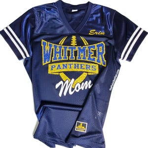 Può includere: Una maglia blu navy con bordini bianchi e dorati. La maglia ha le parole "Whitmer Panthers" in glitter dorato con una grafica di un pallone da calcio. La parola "Mamma" è in glitter bianco e il nome "Erin" è in glitter dorato.