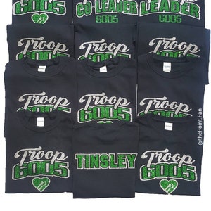K&ouml;nnte beinhalten: Zw&ouml;lf schwarze T-Shirts mit gr&uuml;nem und silbernem Glitzertext. Der Text auf den Shirts lautet "Troop 6005", "Troop 6005 Co-Leader", "Troop 6005 Leader", "Troop Tinsley" und "Troop 6005" mit einem Herzen.