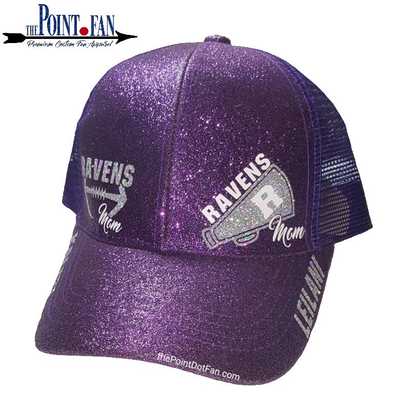 May include: A purple glitter trucker hat with a mesh back. The hat features the text "RAVENS Mom" in silver glitter, along with a football and a megaphone graphic. The name "LEILANI" is printed on the brim. The Point Fan logo is visible.