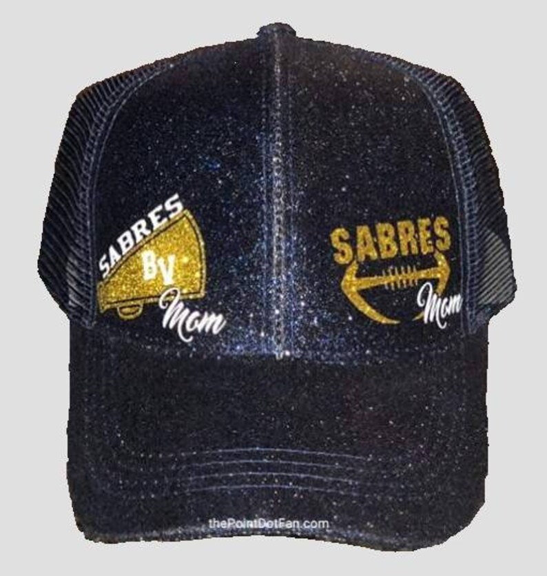 Pu&ograve; includere: Un cappello da camionista nero e oro con glitter con le parole "Sabres Mom" e una grafica di pallone da calcio e megafono dorati.