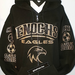 May include: Black zip-up hoodie with gold glitter lettering that reads "ENOCHS EAGLES" and "CLASS OF 2019". The hoodie has a gold glitter eagle on the front and gold glitter sports icons on the sleeves.