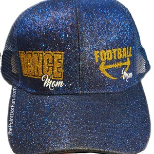May include: A navy blue glitter trucker hat with the words "DANCE Mom" and "FOOTBALL Mom" in gold lettering. The hat has a mesh back and a curved brim. The website address thePointDotFan.com is printed on the side of the hat.