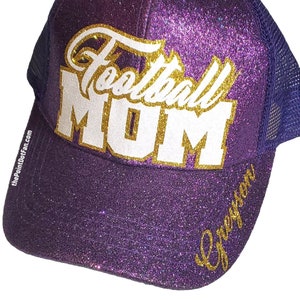 Pode incluir: Um bon&eacute; trucker roxo com glitter apresentando o texto Football MOM em branco e dourado no painel frontal. A aba exibe o nome Greyson em letra dourada e a lateral inclui o pequeno texto branco thePointDotFan.com.