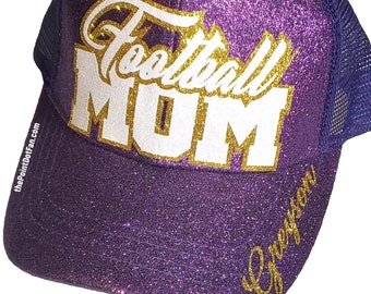 May include: A purple glitter trucker hat featuring the white and gold text Football MOM on the front panel. The brim displays the name Greyson in gold script, and the side of the brim includes the small white text thePointDotFan.com.
