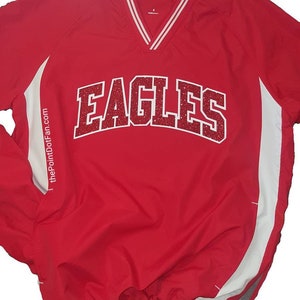 May include: A red long-sleeve pullover windbreaker with a V-neck collar featuring white stripes. The front displays the word EAGLES in red glitter lettering with a white outline. The text thePointDotFan.com is printed vertically on the side.