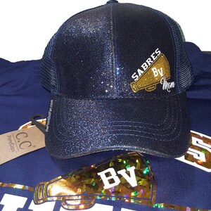 Pu&ograve; includere: Un cappello da camionista blu navy con glitter con un megafono dorato con glitter e il testo "SABRES BV Mom".