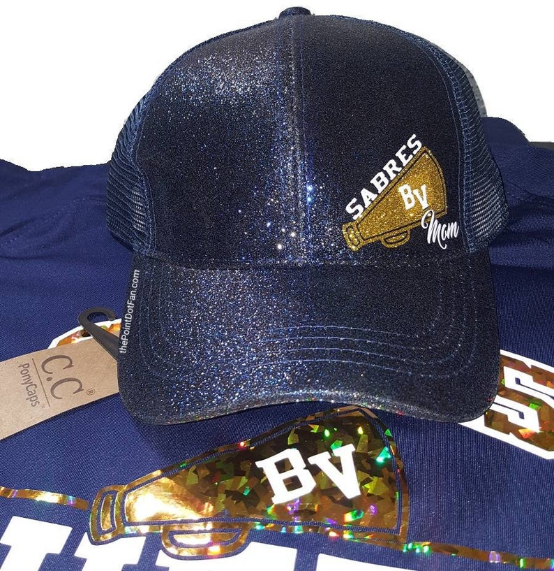 Pu&ograve; includere: Un cappello da camionista blu navy con glitter con un megafono dorato con glitter e il testo "SABRES BV Mom".