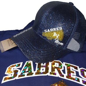 May include: A navy blue glitter baseball cap with a mesh back. The cap features the text "SABRES By Mom" in gold and white. A navy blue shirt with the word "SABRES" in holographic lettering is also visible.