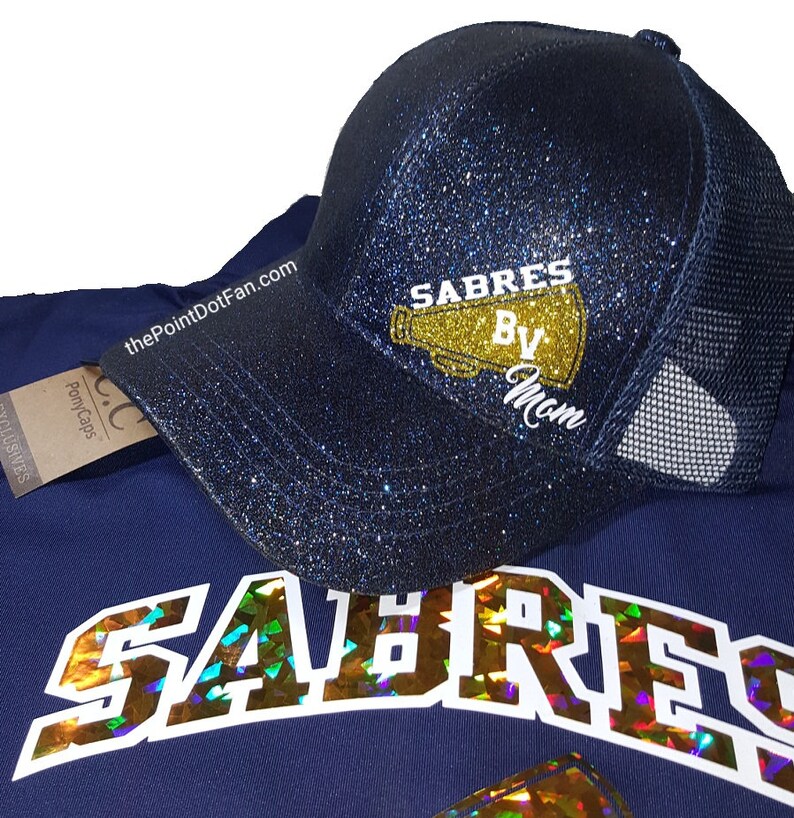 May include: A navy blue glitter baseball cap with a mesh back. The cap features the text "SABRES By Mom" in gold and white. A navy blue shirt with the word "SABRES" in holographic lettering is also visible.