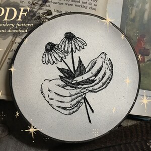 May include: Black and white embroidery design in a hoop. The image shows two flowers held by hands. Text on the image reads "PDF embroidery pattern instant download."