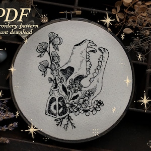 May include: A black and white embroidery pattern featuring a skull with floral elements and the text "PDF embroidery pattern instant download" in a round hoop. The design includes a skull, flowers, and a small bottle.