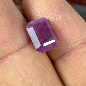 以下が含まれることがあります： 鮮やかな紫色の色合いを持つ、ファセットカットされた長方形の宝石。この石は、鋭いエッジと光を反射する明確な幾何学的なファセットを備えた研磨された表面を持ち、半透明の品質と深く飽和したバイオレットカラーを示しています。