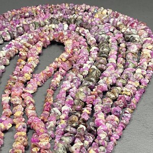 Könnte beinhalten: Stränge aus rohen Rubin-Edelsteinperlen in verschiedenen Rosa-, Lila- und Grautönen. Die unregelmäßig geformten Perlen sind aufgefädelt und erzeugen ein strukturiertes und natürliches Aussehen. Ideal für die Schmuckherstellung.