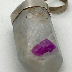 May include: A silver ring with a clear crystal pendant containing a pink ruby. The crystal is faceted and has a rough texture.