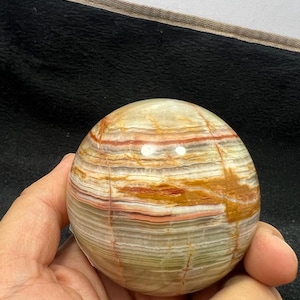 Puede incluir: Una esfera de ónix pulida, redonda y lisa, con un patrón en capas de rayas blancas, verdes, naranjas y rojas.