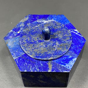Op de afbeelding: Zeshoekige lapis lazuli doos met een cirkelvormig deksel en een kleine, cilindrische handgreep. De doos is diepblauw met gouden vlekken, het deksel is lichter van kleur. Een decoratief item.