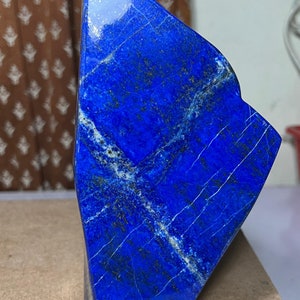 Puede incluir: Una piedra grande de lapislázuli de color azul oscuro, de corte tosco, con vetas blancas y motas de pirita dorada. La piedra está pulida y tiene una superficie lisa y brillante.