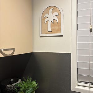 May include: A white palm tree silhouette inside a white arched frame, mounted on a light wood panel. The wall art is displayed on a cream-colored wall. A small potted plant and a mortar and pestle are on the counter below.