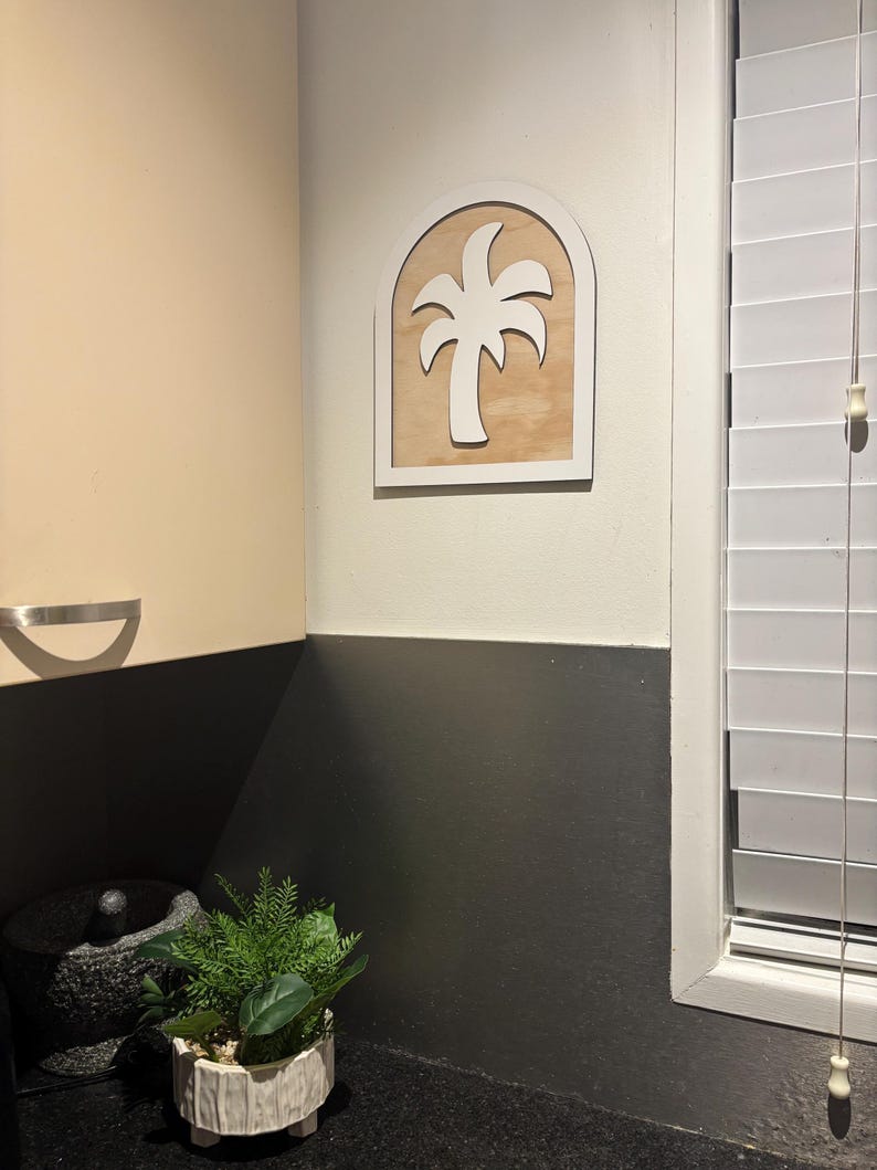 May include: A white palm tree silhouette inside a white arched frame, mounted on a light wood panel. The wall art is displayed on a cream-colored wall. A small potted plant and a mortar and pestle are on the counter below.