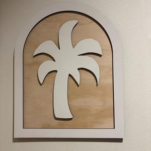 May include: A white palm tree silhouette is centered on a light wood grain background, framed by a white arch. The wooden wall art has a tropical theme and is mounted on a white wall.