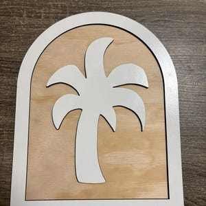 May include: A wooden sign featuring a white palm tree silhouette within a white arched frame. The background is a light brown wood grain. The sign is designed for wall decor.