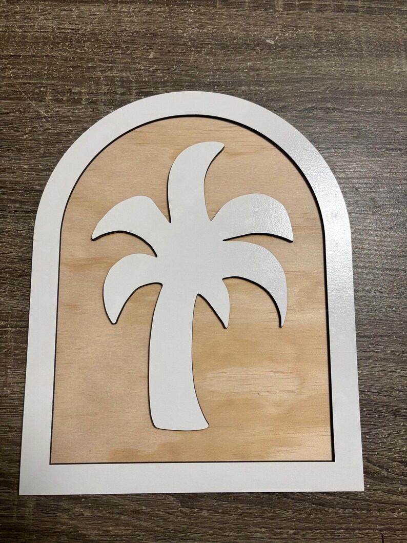 May include: A wooden sign featuring a white palm tree silhouette within a white arched frame. The background is a light brown wood grain. The sign is designed for wall decor.