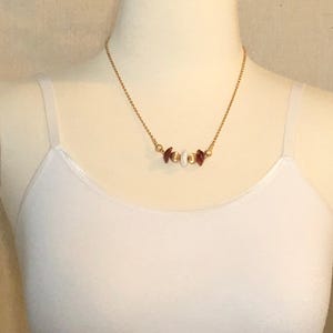 May include: A gold chain necklace featuring a series of small, alternating white and burgundy beads. The necklace is displayed on a white mannequin torso, highlighting the jewellery's design and colour contrast.