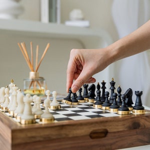 Op de afbeelding: Een houten schaakspel met zwarte en witte stukken. De zwarte stukken hebben goudkleurige accenten. Een hand beweegt een zwarte pion op het schaakbord. Het schaakbord staat op een houten kist.