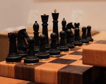 Può includere: Una scacchiera di legno con pezzi degli scacchi neri disposti sulla scacchiera. La scacchiera è fatta di legno chiaro e scuro e ha una finitura lucida.