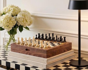 May include: A wooden chess set with cream and black pieces featuring gold-tone bases, resting on two stacked books titled Chess Bois and The Refined Dogs. The set is placed on a black and white checkered surface next to a black lamp and white flowers.