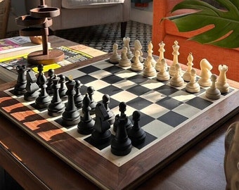 Könnte beinhalten: Ein Schachspiel mit schwarzen und weißen Figuren auf einem Schachbrett angeordnet. Das Brett steht auf einem dunklen Holztisch. Die Schachfiguren sind aus hellem Material gefertigt. Das Brett ist mit einem dunklen Holzrahmen eingefasst.