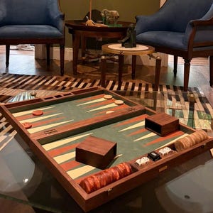May include: A backgammon board with a green playing surface, orange and white triangles, and wooden game pieces. The board is open on a glass-topped table in a room with blue armchairs and a painting.