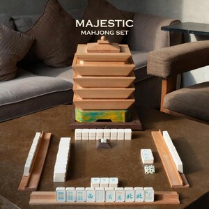 May include: A wooden Mahjong set with the text "MAJESTIC MAH JONG SET". The set includes a stackable wooden box, wooden tile holders, and white tiles with symbols and numbers. The box has a green and yellow painted base.