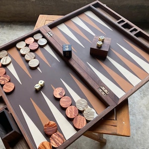 May include: A wooden backgammon board with brown, tan, and white triangular points. The board is open, revealing wooden game pieces in shades of brown and tan, along with dice. The game is set on a wooden table.