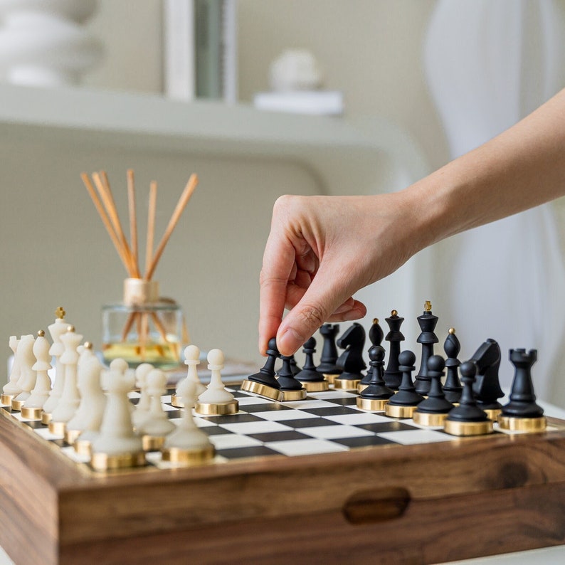 Op de afbeelding: Een houten schaakspel met zwarte en witte stukken. De zwarte stukken hebben goudkleurige accenten. Een hand beweegt een zwarte pion op het schaakbord. Het schaakbord staat op een houten kist.