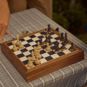 Op de afbeelding: Een houten schaakspel met zwarte en cr&egrave;mekleurige stukken op een schaakbord. Het bord staat op een grijs-witte geruite tafelkleed. Sommige stukken zijn omvergeworpen, wat suggereert dat er een spel aan de gang is.