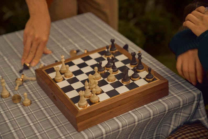 Op de afbeelding: Een houten schaakspel met zwarte en cr&egrave;mekleurige stukken op een schaakbord. Het bord staat op een grijs-witte geruite tafelkleed. Sommige stukken zijn omvergeworpen, wat suggereert dat er een spel aan de gang is.