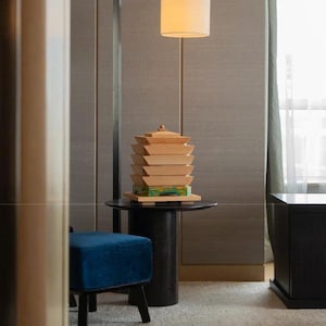 May include: A modern interior scene featuring a wooden pagoda sculpture on a black side table, illuminated by a cylindrical lampshade. A blue velvet ottoman and a dark cabinet are also visible.