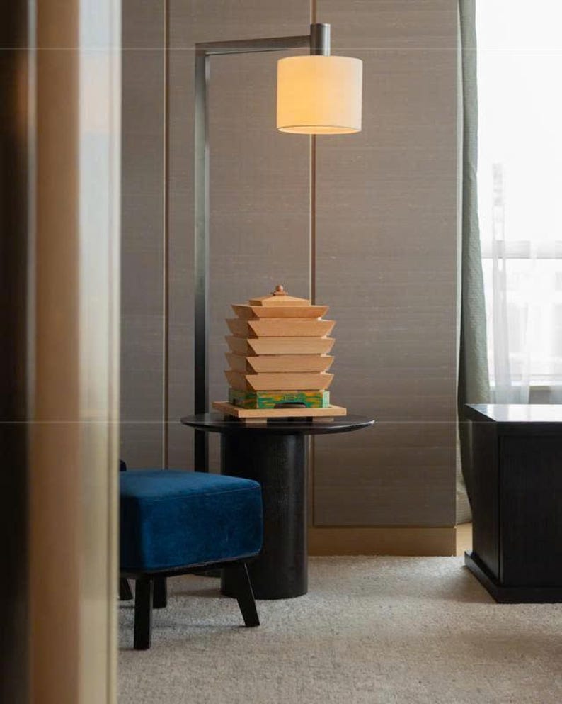 May include: A modern interior scene featuring a wooden pagoda sculpture on a black side table, illuminated by a cylindrical lampshade. A blue velvet ottoman and a dark cabinet are also visible.