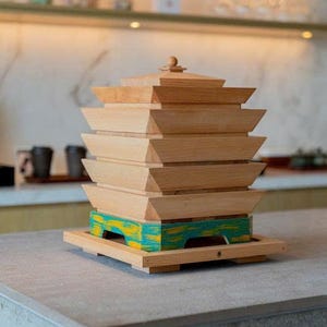 May include: A wooden decorative object with a pagoda-like design. The natural wood structure features multiple stacked layers, with a base painted in teal and yellow. The object sits on a gray countertop.