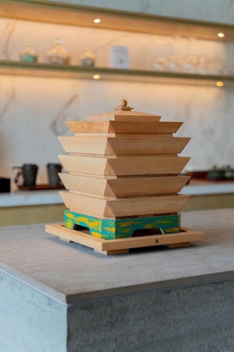 May include: A wooden decorative object with a pagoda-like design. The natural wood structure features multiple stacked layers, with a base painted in teal and yellow. The object sits on a gray countertop.