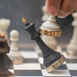Op de afbeelding: Een zwarte schaakkoning met gouden accenten wordt omvergeworpen door een wit schaakstuk. De schaakstukken staan op een zwart-wit geblokt bord. Andere schaakstukken zijn op de achtergrond te zien.